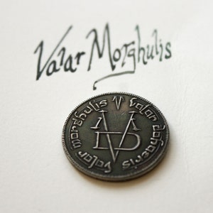 Op de afbeelding: Een zilveren munt met de inscriptie "Valar Morghulis" in een cirkelvormige patroon rond de rand. Het midden van de munt heeft een gestileerde "V" met twee kruisende zwaarden.
