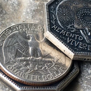 May include: A silver US quarter dollar coin with an eagle and the words "United States of America" and "Quarter Dollar" on it. The coin is on top of a silver coin with the words "Memento Vivere" on it.