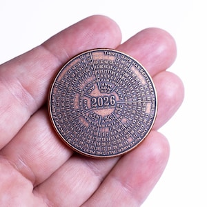 May include: A bronze-colored, circular calendar coin for the year 2026, held in a hand. The coin features months, dates, and the text "THIRTY DAYS HAS SEPTEMBER, APRIL JUNE, AND NOVEMBER. ALL THE REST HAVE 31 EXCEPT FEBRUARY".