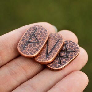 May include: Four copper-colored metal tokens with different Norse runes engraved on them. The runes are in a black color and are visible on the front of each token.