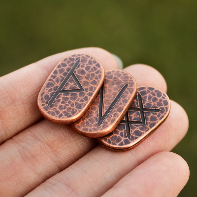 May include: Four copper-colored metal tokens with different Norse runes engraved on them. The runes are in a black color and are visible on the front of each token.