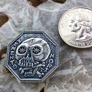May include: A silver-toned octagonal coin with a skull design and the words "Memento Mori" inscribed around the edge.