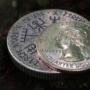 May include: A silver quarter dollar coin with the inscription "United States of America" and "Liberty" on the front, and "Quarter Dollar" on the back, is placed on top of a silver coin with a design of symbols and the inscription "#MR.FOX.TRAMA".