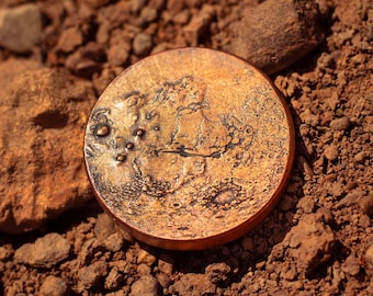 May include: A close-up of a copper coin with a textured surface and a design that resembles the moon's surface. The coin is lying on a bed of reddish-brown dirt.