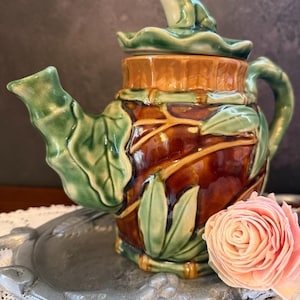 Op de afbeelding: Een keramische theepot met een bruin lichaam en groene bladaccenten, voorzien van een deksel met daarop een klein groen kikkerbeeldje. De theepot staat op een zilverkleurig metalen dienblad naast een enkele, lichtroze kunstbloem.