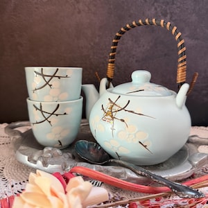 May include: A light blue ceramic teapot with a curved handle wrapped in dark brown material, accompanied by two matching tea cups. The set features a painted design of white blossoms and dark brown branches, with gold-coloured Japanese text.