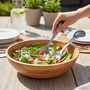 May include: A wooden salad bowl filled with a fresh salad of spinach, tomatoes, cucumbers, and red onions. Two silver serving spoons with seashell-shaped handles are in the bowl. The salad is on a wooden table.
