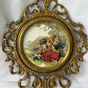May include: Ornate, gold-toned metal frame with a round, hand-painted porcelain insert. The insert depicts a romantic scene with figures in period clothing, a dog, and a landscape. The frame has a decorative, openwork design.