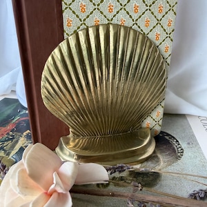 May include: A polished brass bookend shaped like a large scallop shell with prominent vertical ridges and a smooth, curved base. The metallic surface reflects light, highlighting the shell's fan-like structure and its solid, sturdy construction.