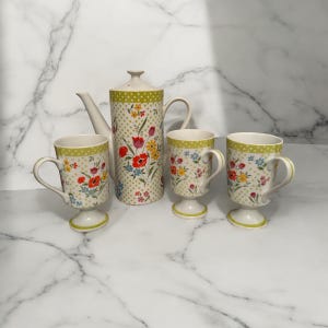 Puede incluir: Un juego de cuatro tazas de cerámica con estampado floral y una tetera a juego. Las tazas tienen un estampado de lunares verdes y blancos con un diseño floral. La tetera tiene un diseño a juego y una tapa.