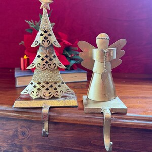 May include: Two gold-toned metal stocking holders. One features a cutout Christmas tree design topped with a star and glitter accents. The other is a stylised angel figure with wings and a small heart charm hanging from a chain.