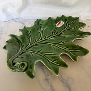 May include: A green ceramic serving dish shaped like an oak leaf with a glossy finish and raised vein details. A small oval sticker on the surface reads Hand Painted by Artisans. The dish features a curled stem at the base and jagged edges.