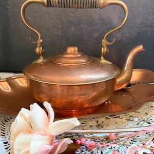May include: A copper teapot with a rounded body, a curved spout, and a brass handle featuring a wrapped textured grip. The teapot rests on a matching oval copper tray. The surface of the metal shows a warm, polished finish with subtle patina.