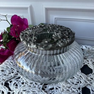 May include: A clear glass jar with a ribbed design and a decorative silver lid. The jar is round and sits on a white lace doily. Bright pink flowers are visible in the background.