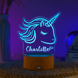 以下が含まれることがあります： アクリルに「Charlotte」と刻まれた青いLEDユニコーンのナイトライト。ユニコーンのデザインは木製のベースの上にあります。背景は紫と青のグラデーションです。