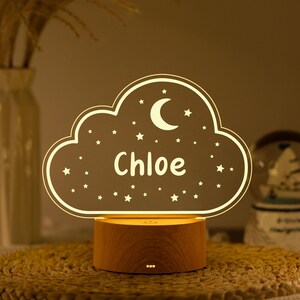 May include: A personalized night light shaped like a cloud, featuring the name "Chloe" in white script. The cloud design includes a crescent moon and stars. It sits on a wooden base, creating a warm, ambient glow.