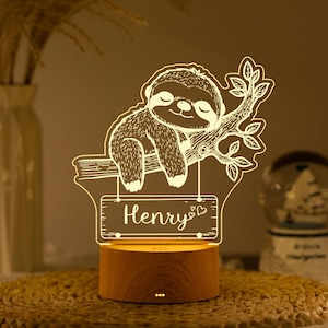 Può includere: Luce notturna decorativa con un design di bradipo addormentato su un ramo, con il nome "Henry" su un cartello. La lampada ha una base in legno ed emette una calda luce. Il bradipo è dettagliato con linee incise.