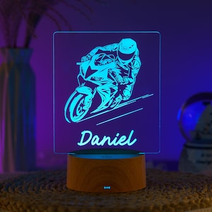 Puede incluir: Una luz de noche de motocicleta azul neón con el nombre "Daniel" en escritura blanca. La luz presenta una ilustración detallada de un corredor de motocicletas en una pista, sobre una base de madera.