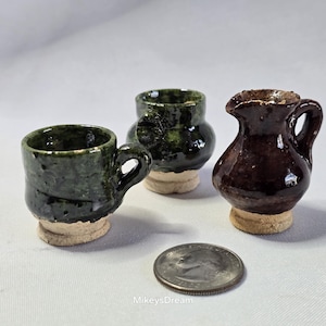 May include: Three miniature ceramic vessels: a dark green mug with a handle, a dark green pot, and a brown pitcher. Each sits on a light tan base. A quarter is next to the vessels for scale. The vessels are handmade.