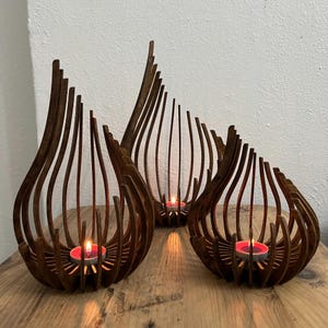 May include: Three wooden candle holders with a teardrop design. The holders are made of thin, curved wooden slats, creating an open, airy structure. Each holder contains a lit red tea light candle, casting a warm glow. The holders are on a wooden surface.
