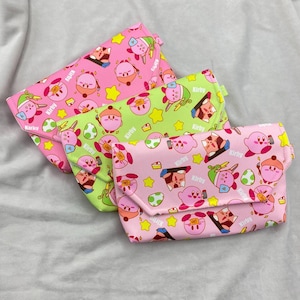 Puede incluir: Tres bolsos estampados de colores con un diseño de personajes de dibujos animados repetidos. Los bolsos son rosas, verdes y amarillos, con cierre de solapa. El diseño incluye estrellas, huevos y la palabra "Kirby".