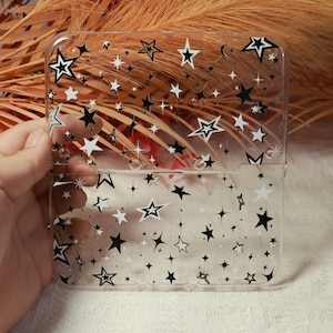 May include: Transparent phone case featuring a pattern of black and white stars. The stars vary in size and are scattered across the case. The case has a rectangular shape and a glossy finish, creating a celestial theme.