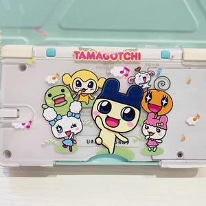 May include: A clear plastic Tamagotchi device featuring colorful cartoon characters. The device has a white frame with the word "TAMAGOTCHI" in pink. The characters include a monkey, a duck, and other animated figures.