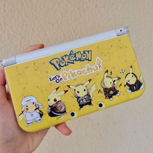 Puede incluir: Consola de juegos portátil Nintendo 3DS amarilla con personajes de Pikachu con diferentes atuendos. La consola tiene el texto "Pokémon Let's Go Pikachu!" e ilustraciones de Pikachu. La consola se sostiene en una mano.