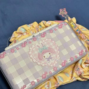 Könnte beinhalten: Eine Nintendo 3DS-Hülle mit lila und weißem Karomuster, die einen Cartoon-Hasencharakter in einem herzförmigen Design zeigt, umgeben von rosa Rosen. Ein gelber Kordelzugbeutel und ein dekorativer Zauberstab sind ebenfalls sichtbar.