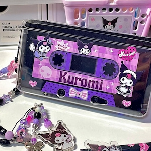 May include: A clear phone case in the shape of a cassette tape, with a purple and pink Kuromi design. It includes a beaded charm and a matching pen. The text "Kuromi" and "You all rock!" are displayed on the case.