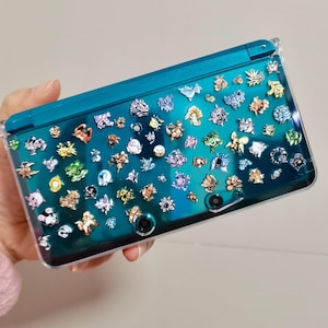 Könnte beinhalten: Eine türkisfarbene Nintendo 3DS-Handheld-Konsole mit einer transparenten Schutzhülle. Die Hülle ist mit bunten Aufklebern von Zeichentrickfiguren bedeckt. Die Konsole wird in der Hand einer Person gehalten.
