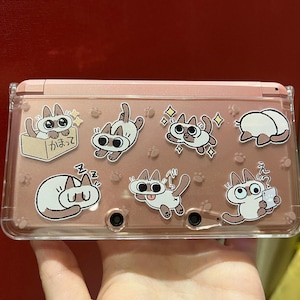 Può includere: Una custodia rosa per Nintendo 3DS decorata con adesivi di gatti dei cartoni animati. Gli adesivi raffigurano gatti in varie pose, come dormire, in scatole e con una tazza. La custodia ha una copertura in plastica trasparente e pulsanti neri.