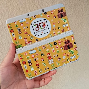 Puede incluir: Consola de juegos portátil Nintendo 3DS amarilla con un diseño del 30 aniversario de Super Mario Bros. La consola abierta muestra un patrón de personajes de Mario, champiñones y gráficos de castillo.