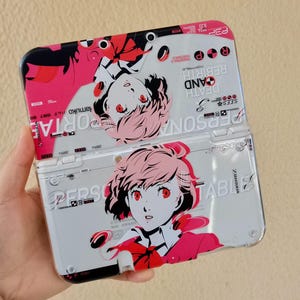 May include: A white Nintendo 3DS case featuring a pink and red anime-style illustration of a character with pink hair and red eyes. The case has the words "PERSONA" and "REBIRTH" printed on it.