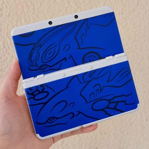 May include: A blue and white Nintendo 3DS handheld gaming console. The console features a blue design with a black illustration of a creature. The console is open, revealing the screen and buttons.