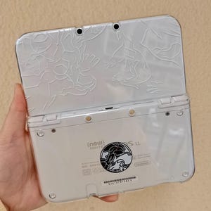 Puede incluir: Consola de juegos portátil Nintendo New 3DS LL blanca con una funda protectora transparente. La cubierta superior presenta un diseño en relieve blanco. La parte inferior tiene una pegatina circular en blanco y negro.