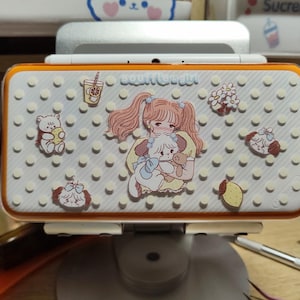 May include: A white and orange handheld gaming device with a decorative cover featuring a cartoon girl holding a puppy, surrounded by other cute illustrations. The device is on a stand.