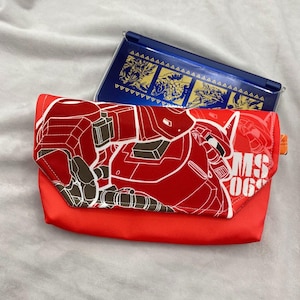 May include: A red clutch with a white and brown graphic of a robot, with the text "MS 06". A blue handheld gaming device with gold graphics is behind the clutch. The clutch has an orange tab.