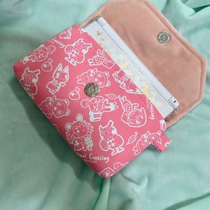 May include: A pink Animal Crossing Nintendo DS case featuring white character illustrations. The case is open, showing a white and gold Nintendo DS. The interior is soft pink fabric, and the case has a magnetic snap closure.