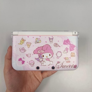 Può includere: Una custodia rosa per Nintendo 3DS con un design di My Melody. La custodia è decorata con vari personaggi dei cartoni animati, fiocchi e la parola "Sweet". La custodia è tenuta in mano.