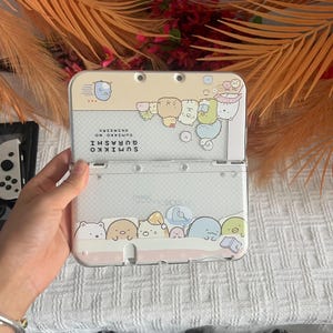 Peut inclure: Une console de jeu portable Nintendo 3DS transparente avec un design Sumikko Gurashi. L'étui présente des personnages de dessins animés aux couleurs pastel, avec le texte "SUMIKKO GURASHI" et du texte japonais. La console est ouverte, révélant l'écran et les boutons.