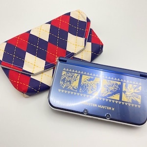 Puede incluir: Una consola de juegos portátil azul con gráficos dorados y el texto "Monster Hunter X" junto a una funda a juego con estampado de rombos rojos, blancos y azules.