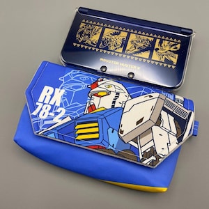 Może przedstawiać: Niebieska i biała kopertówka z motywem RX-78-2 Gundam oraz granatowa konsola Nintendo 3DS. Kopertówka posiada grafikę robota i napis "RX-78-2". 3DS ma złote grafiki i napis "Monster Hunter X".
