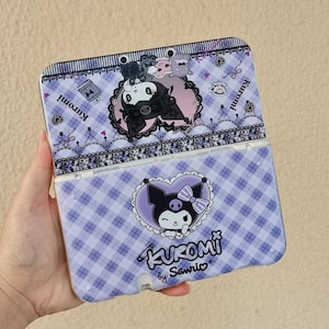 May include: A purple and white checkered Nintendo 3DS case featuring Kuromi characters. The case has a hinged design and is decorated with cartoon graphics, including Kuromi in a heart-shaped design. The text "Kuromi by Sanrio" is also visible.