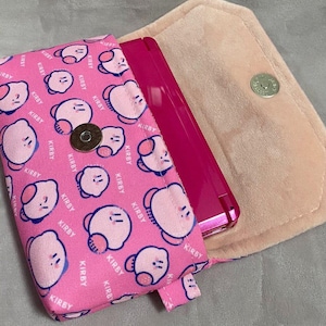 May include: Pink phone case with a repeating Kirby character pattern and the word "KIRBY". The case is open, showing a pink phone. The interior is a soft, light pink material, and the case has a magnetic clasp.