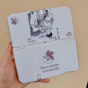 Puede incluir: Consola de juegos portátil Nintendo 3DS blanca con un diseño personalizado con personajes de anime y el texto "NEON GENESIS EVANGELION". La consola está abierta, revelando la pantalla y los botones. El diseño incluye detalles morados y texto japonés.