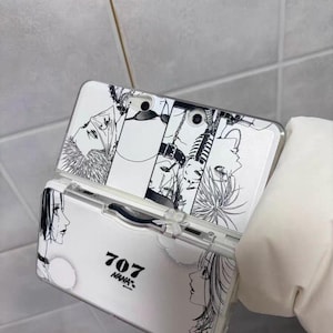 Puede incluir: Funda de teléfono blanca con ilustraciones de personajes en blanco y negro estilo manga. La funda tiene el texto "707 NANA" impreso. La funda está sujeta por una mano enguantada.