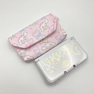 Peut inclure: Une pochette rose clair à motifs avec une fermeture à rabat et un motif de personnage de dessin animé blanc et bleu clair. Une console de jeu portable argentée avec un motif décoratif est à côté de la pochette.