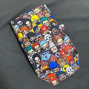 May include: A rectangular wallet with a colourful cartoon character pattern. The design features a variety of characters in different colours, including yellow, red, blue, and green, on a black background. The wallet has a small clasp.