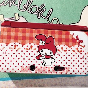 May include: A red and white Nintendo 3DS case with a My Melody design. The case has a red gingham pattern on top, a white polka dot pattern on the bottom, and a My Melody character illustration. The word "MELODY" is printed on the bottom.